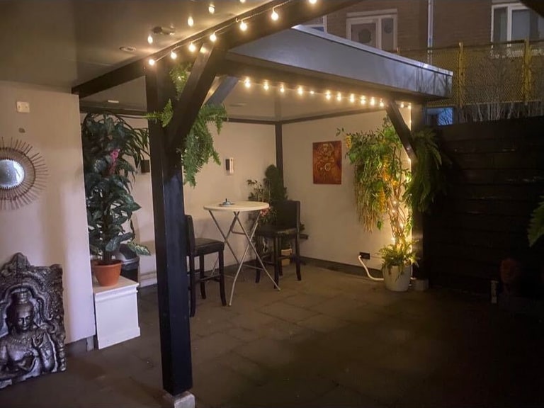 De overkapping in de avond, met lichtsnoeren, een statafel en een paar planten