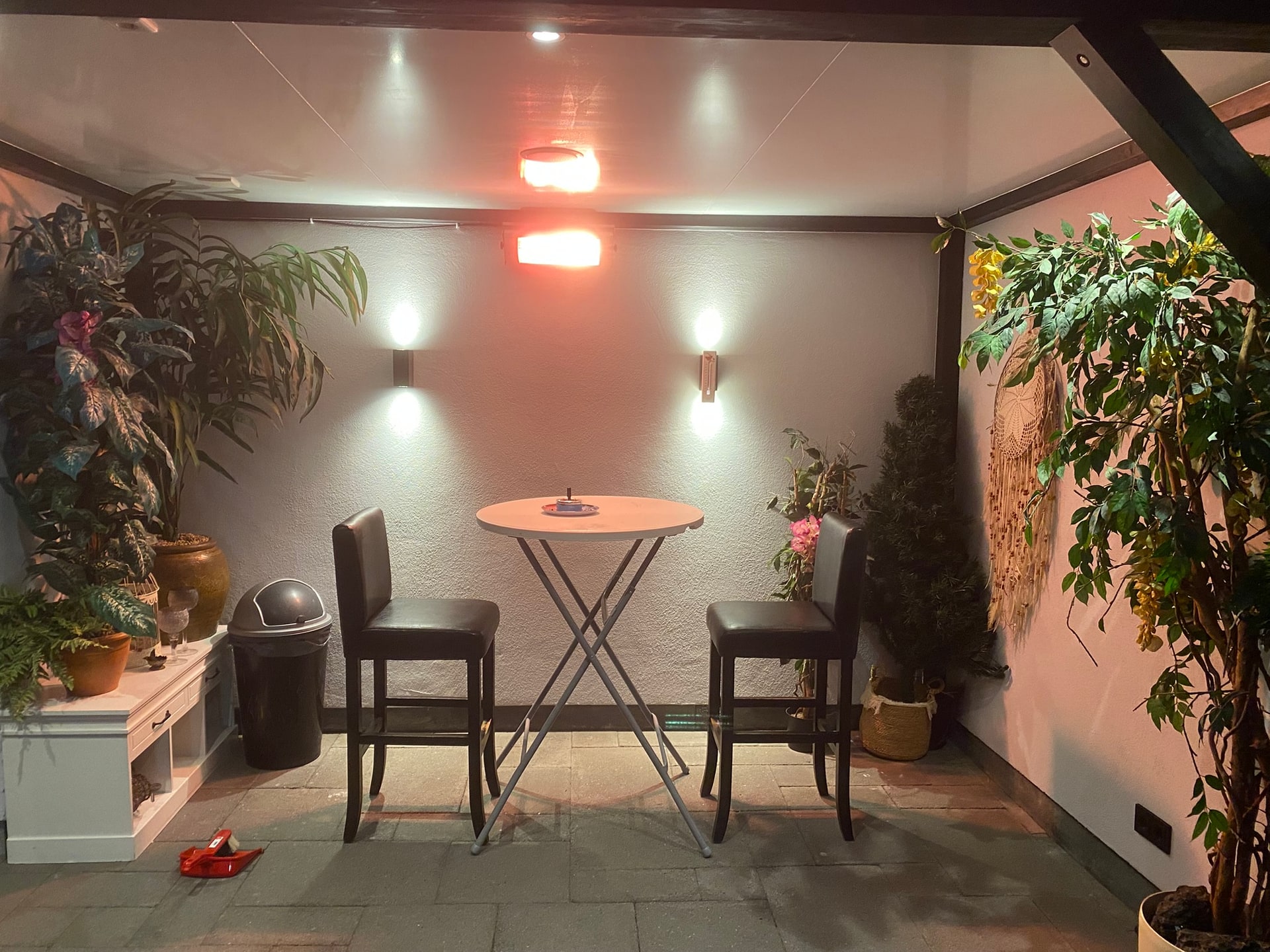 De overkapping in de avond met een warmtelamp aan het plafond, een statafeltje met twee krukken en planten langs de randen