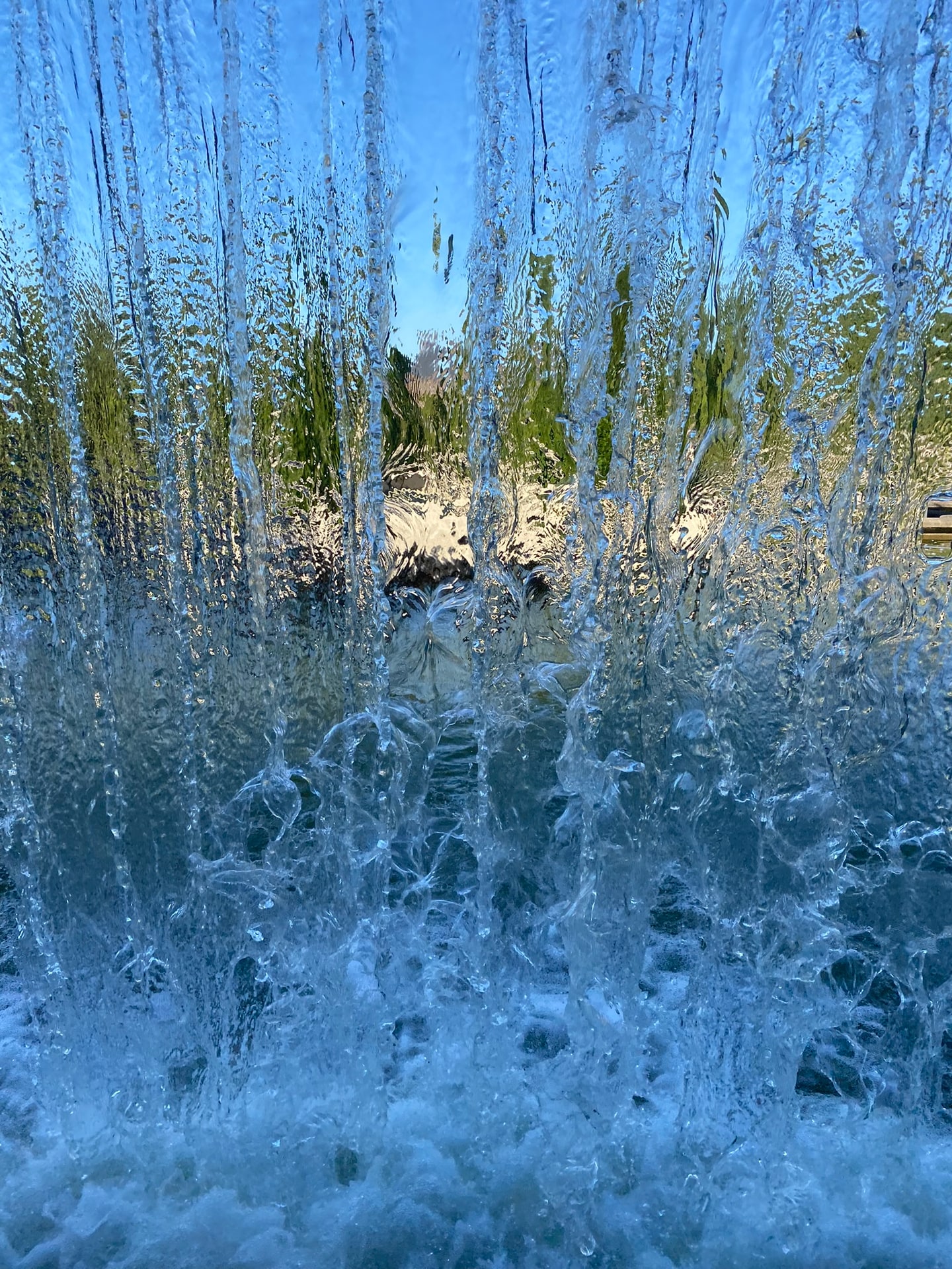 Van dichtbij door een gordijn van vallend fonteinwater, met een wazige tuin met cipressen en blauwe lucht erachter