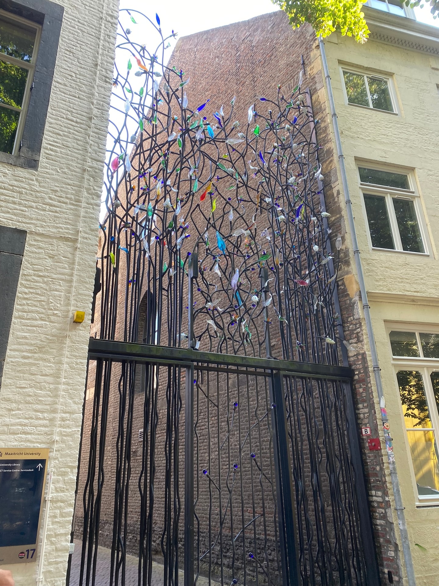 Een hoog smeedijzeren hek in de vorm van vertakte twijgen, versierd met honderden kleurige glazen vogeltjes, tussen twee oude gebouwen