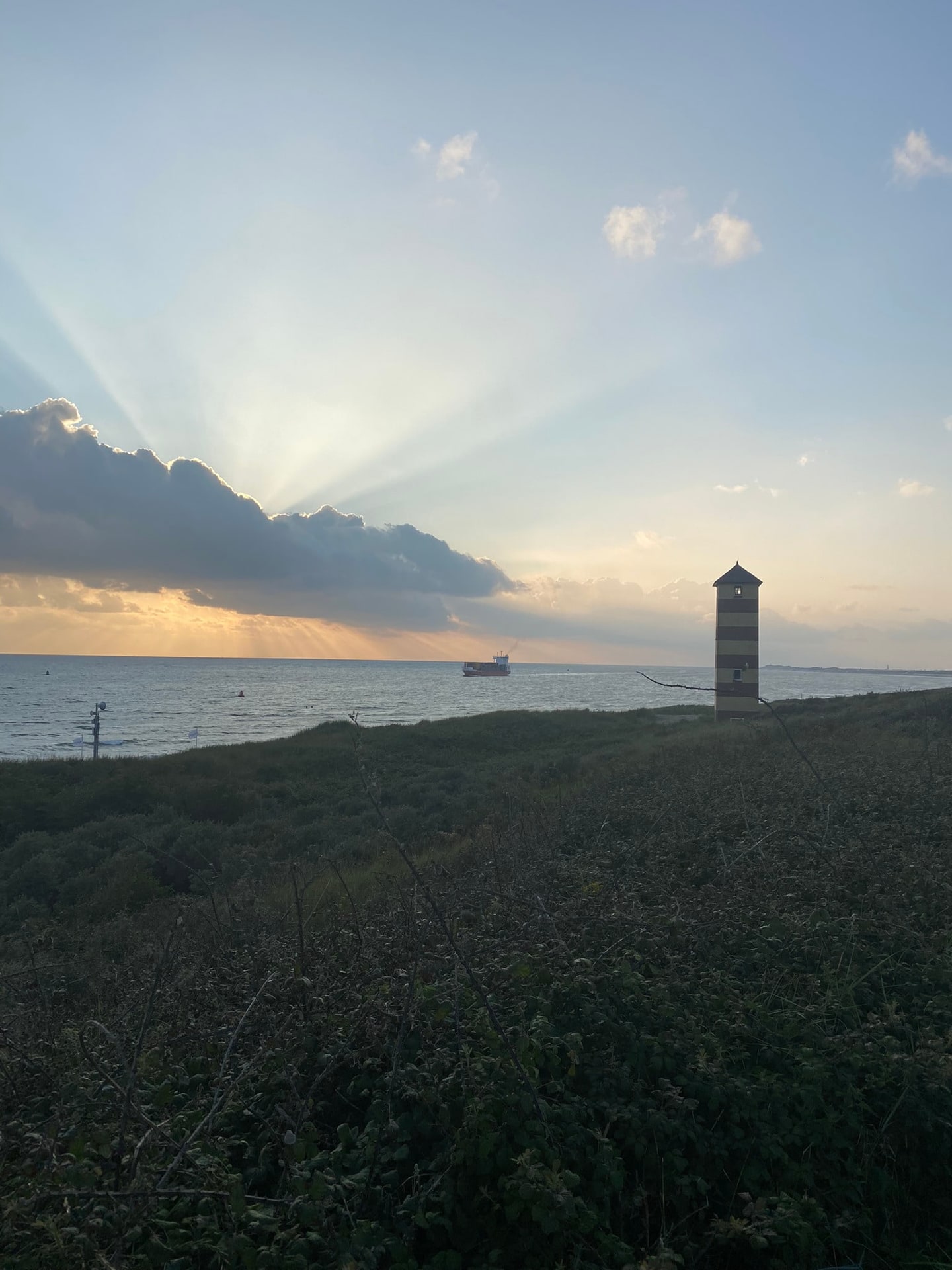 Zonnestralen door de wolken boven zee, met een gestreepte toren in de duinen
