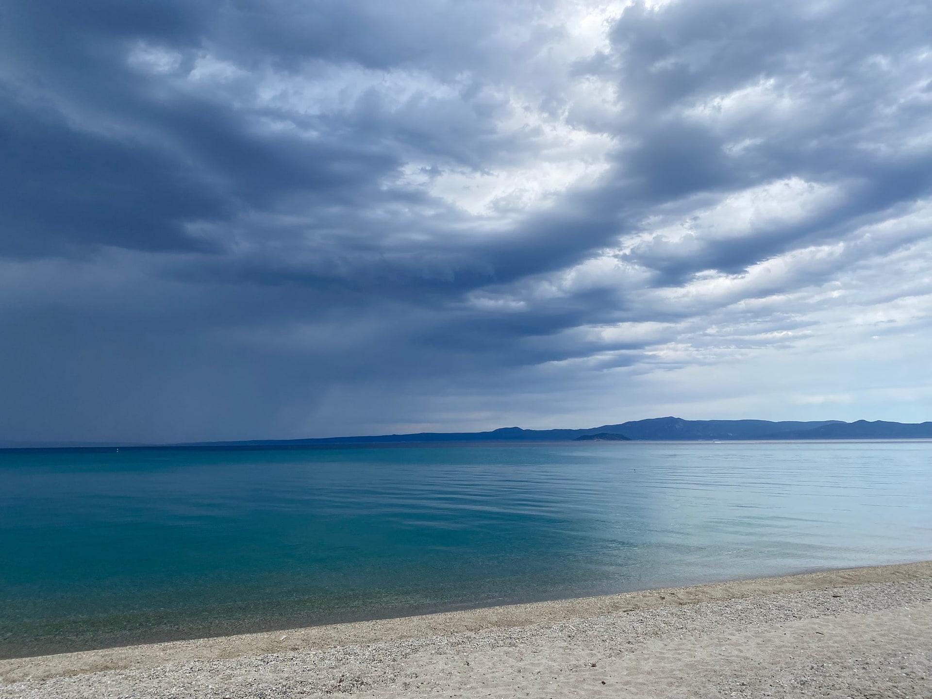 Een zwaar wolkendek met regen in de verte, boven turkooizen zee en een leeg kiezelstrand