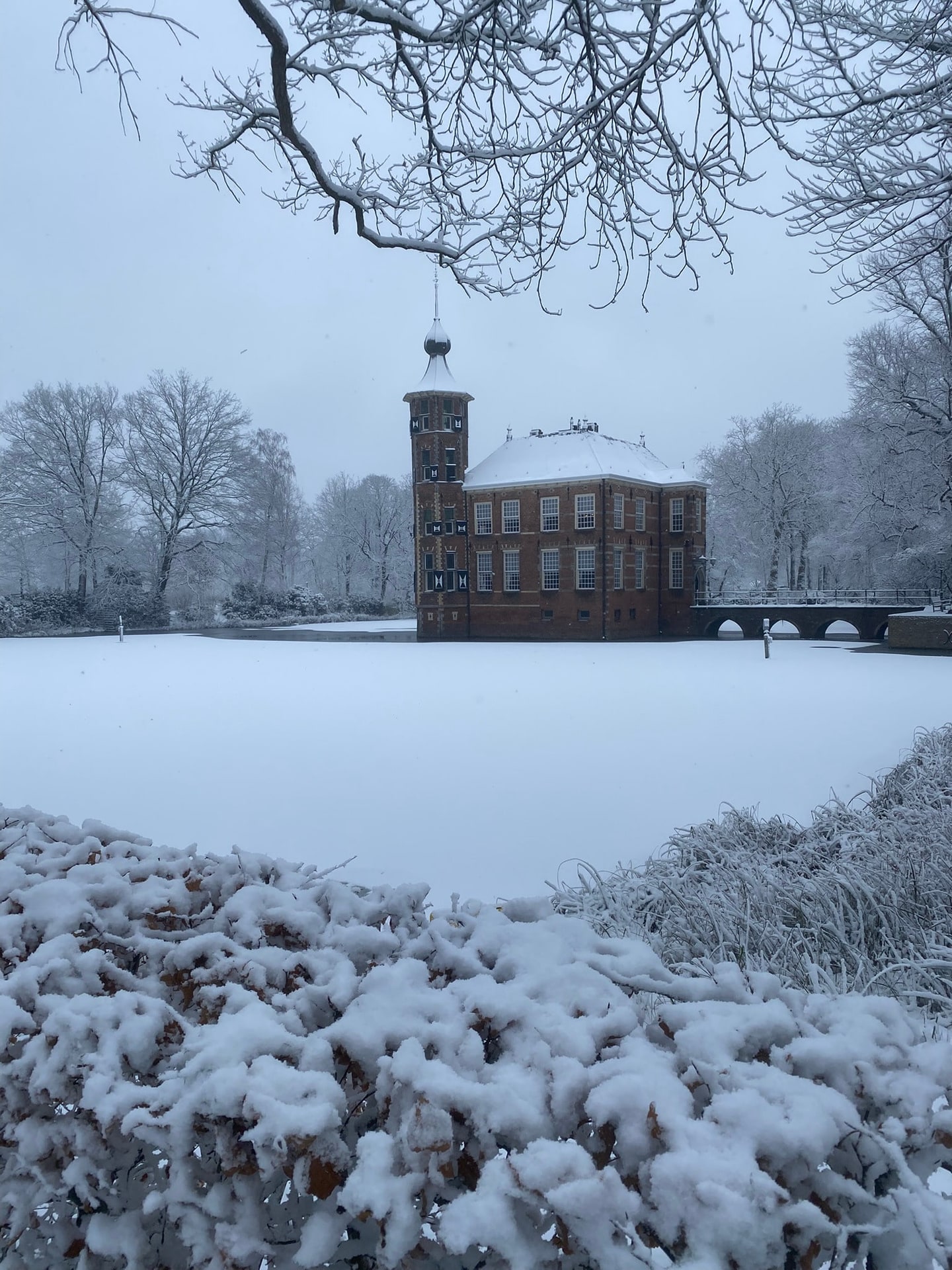 Een landhuis in een besneeuwd park, gezien door kale takken