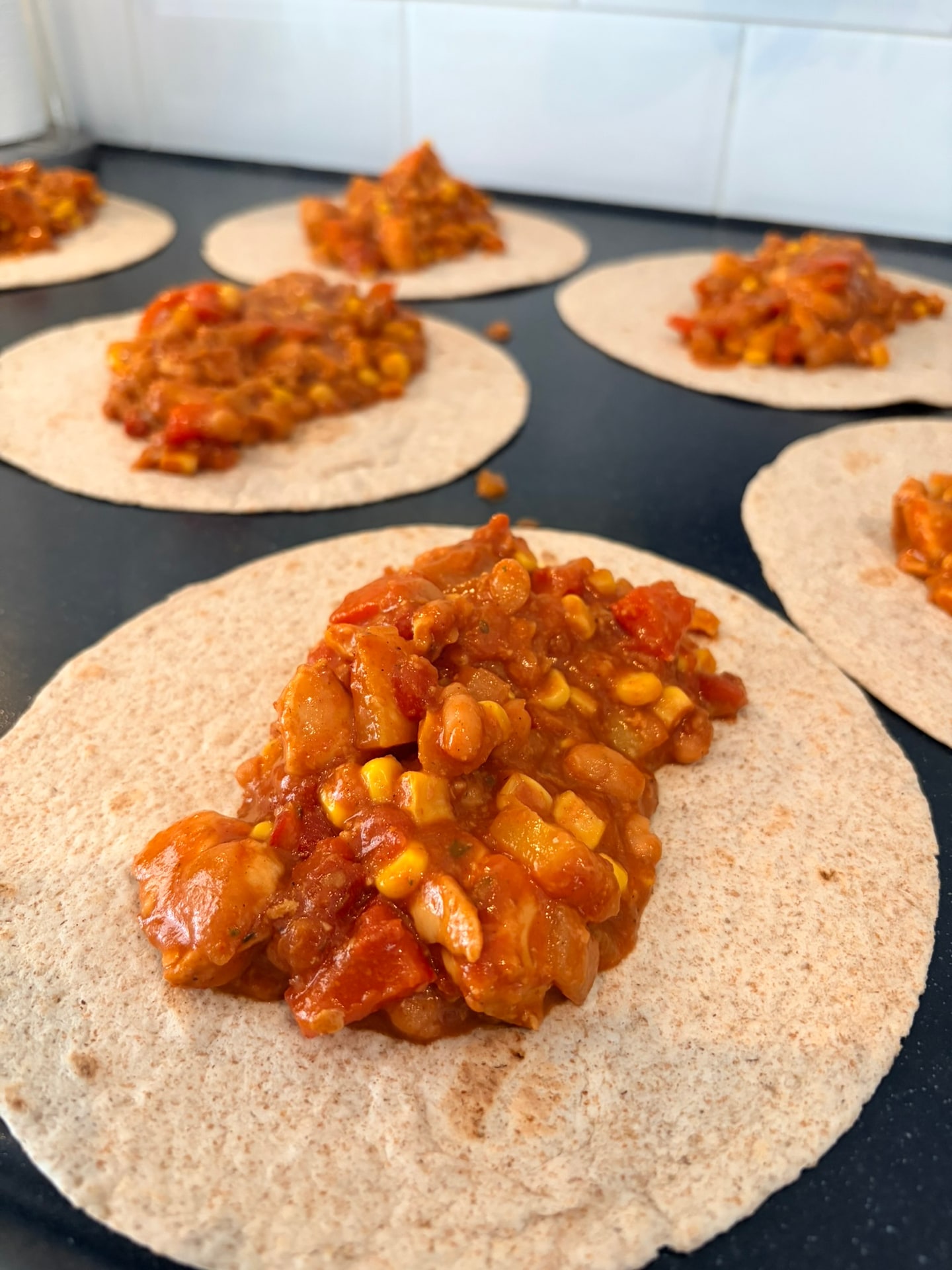 Meerdere volkoren tortilla’s op een donker werkblad, elk met een schep tomatensaus met kip, bonen, mais en paprika erop