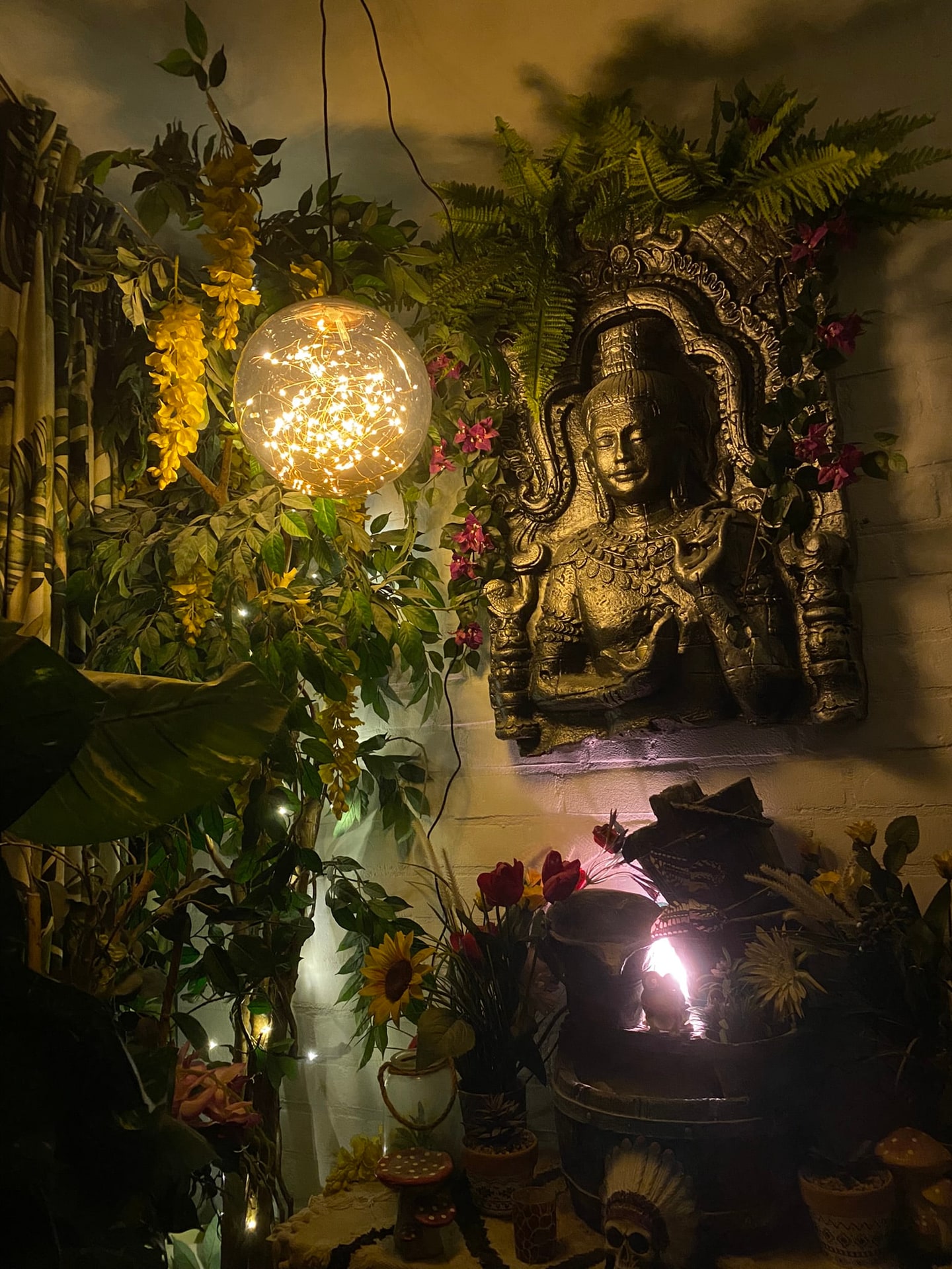 Een verlicht hoekje in de serre in de avond, met een reliëf aan de muur, varens, een fonteintje en een lamp vol lichtjes